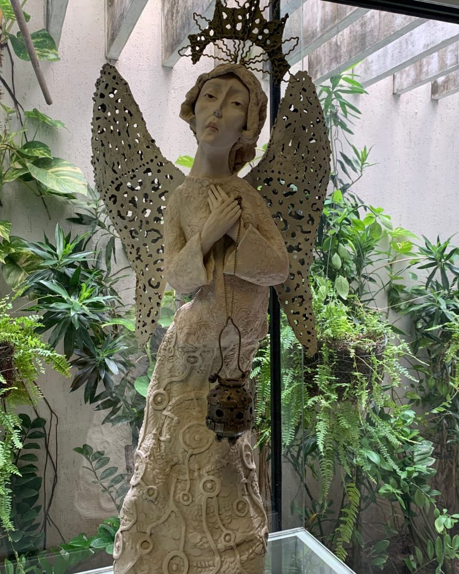 Escultura de anjo com asas de metal vazado e coroa, diante de um jardim interno com plantas.