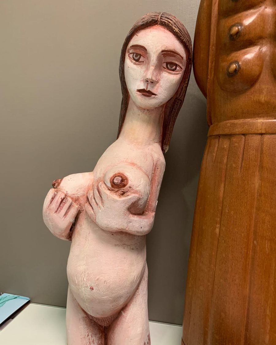 Escultura de figura feminina gestante, pintada em tons de rosa, segurando os seios com as duas mãos.