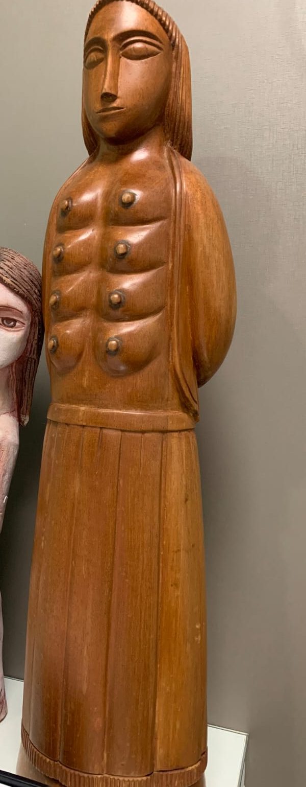 Escultura alta em madeira de uma figura feminina com várias mamas esculpidas no tronco.
